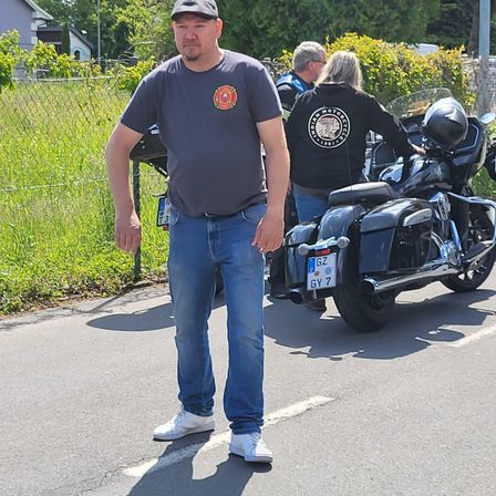 Ein Mann steht neben einem Motorrad und trägt ein Feuerwehr-Patch auf seinem T-Shirt. Dahinter hält eine Frau einen Helm. Das Motorrad hat ein Nummernschild mit den Buchstaben GY 7.