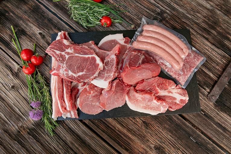 Various cuts of pork, sausages, and fresh herbs on a wooden surface with tomatoes in the background.