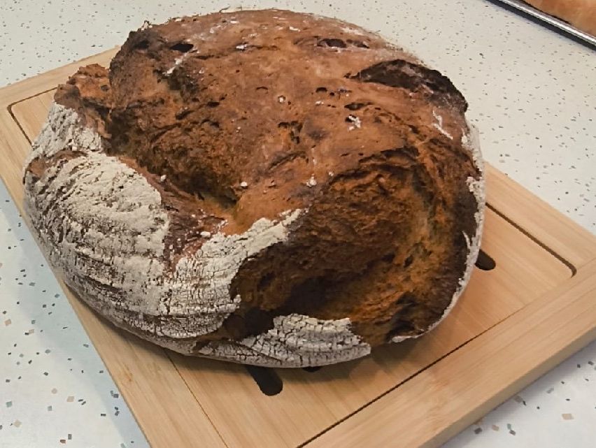 A freshly baked loaf of dark brown bread is shown on a wooden cutting board, placed on a light countertop.