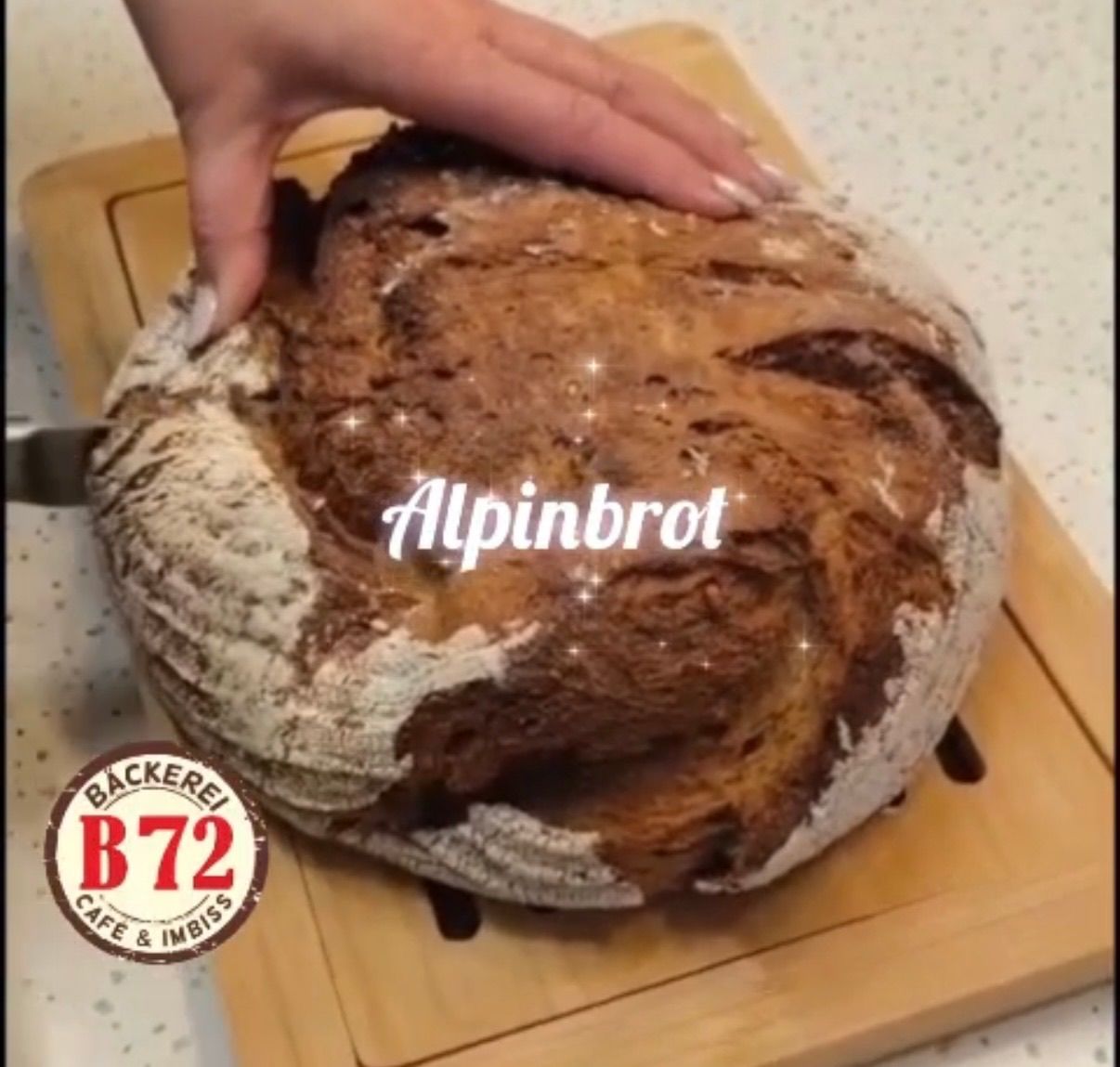 A hand is slicing a whole loaf of bread labeled Alpinbrot on a wooden cutting board. A circular logo with the text Backerei B72 Cafe & Imbiss is in the corner.