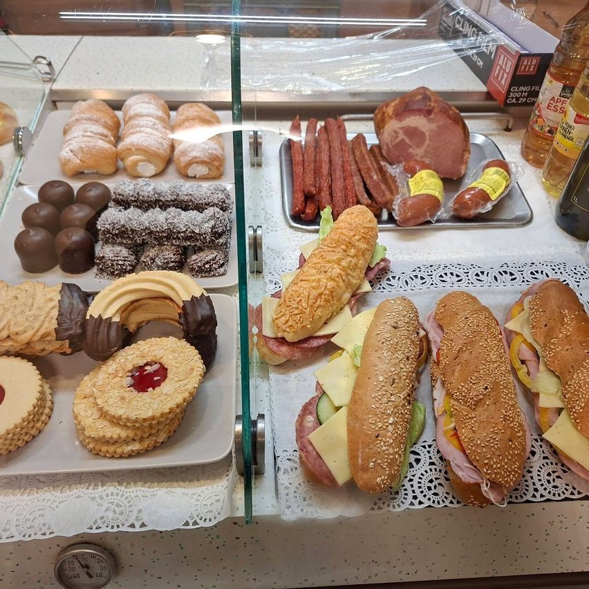 In einer Vitrine sind verschiedene Gebäcksorten, darunter Pralinen und Kekse, neben einer Auswahl an Wurstwaren und Sandwiches ausgestellt. Jedes Produkt ist ordentlich auf Tabletts mit Spitzendeckchen angeordnet.