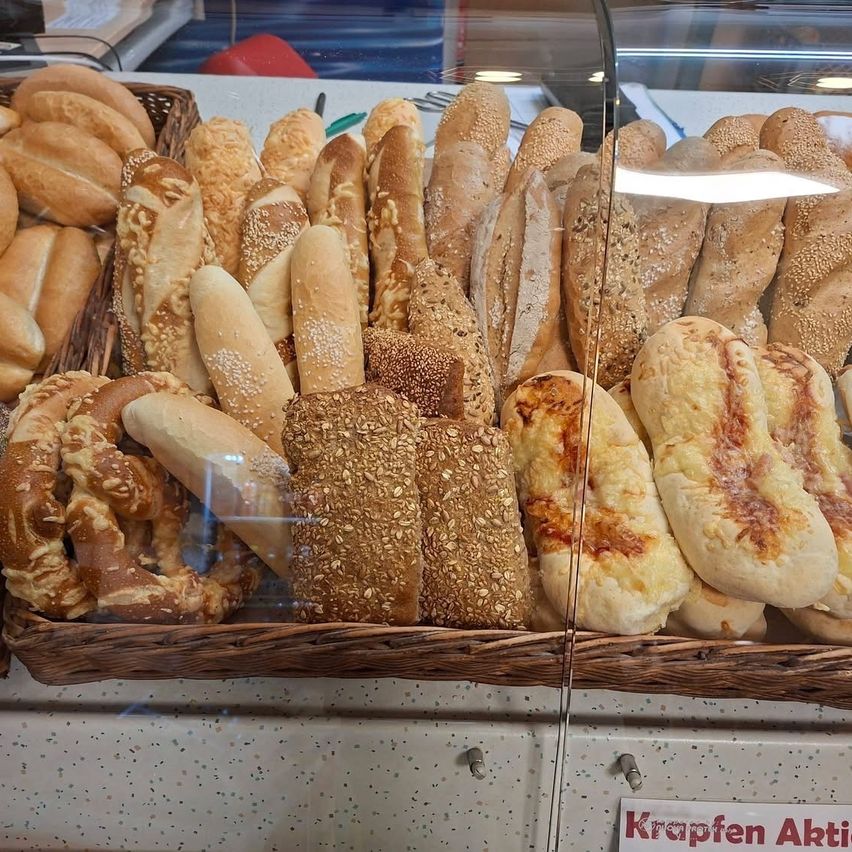 Eine Vitrine mit verschiedenen Brotsorten, darunter Brezeln, Grissini und mit Sesam bestreute Brötchen. Der Tresen ist hinter einer Glasscheibe mit einem roten Schild 'Krappen Aktion'.