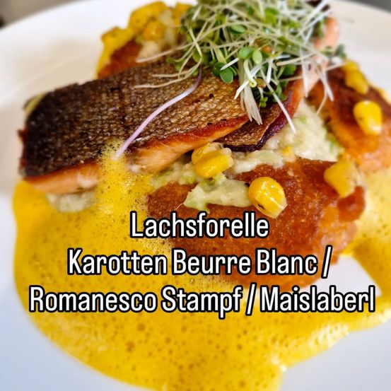 Ein Teller mit Lachsfilet in gelber Sauce, garniert mit Microgreens, Mais und einer Scheibe Brot. Das Gericht wird von einer cremigen Romanesco-Püree begleitet. Auf dem Teller steht 'Lachsforelle Karotten Beurre Blanc / Romanesco Stampf / Maislabel'.
