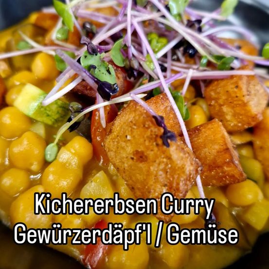 Ein Nahaufnahme eines lebhaften Kichererbsen-Curry-Gerichts mit gerösteten Tofuwürfeln, Avocado und Microgreens-Garnitur. Das Gericht ist in einer schwarzen Schüssel präsentiert, mit dem Text 'Kichererbsen Curry Gewürzerdäpfel / Gemüse' in fetter Schrift.