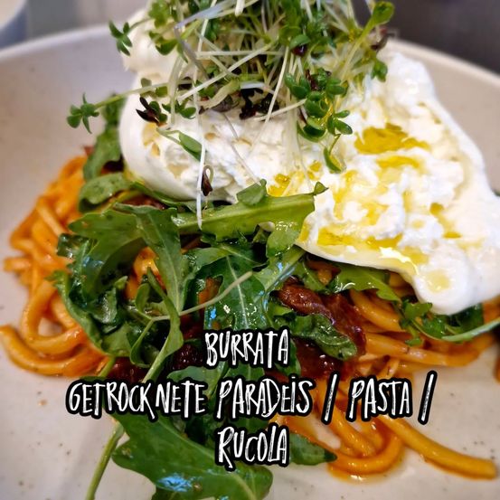 Ein Teller mit Pasta, Burrata-Käse, Rucola und einem Olivenöl-Drizzle obenauf. Das Gericht ist mit Microgreens garniert und hat eine rustikale Präsentation.
