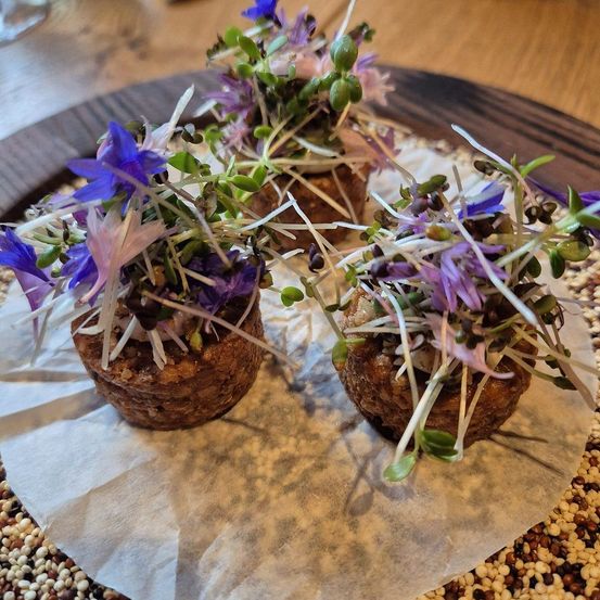 Drei kleine runde Gerichte mit violetten Blumen und grünen Sprossen darauf, auf einem weißen Papier auf einer Holzoberfläche.