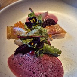 Ein weißer Teller mit einem Gericht, das eine rosarote Rote Bete, grüne Blätter, violette Blumen und ein Stück Fisch mit Kaviar oben drauf zeigt.