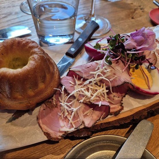 Ein Nahaufnahme eines Holztisches mit geschnittenem Schinken, Käse und einer Blume darauf, einem Brötchen und einem Messer. Es gibt auch ein Glas Wasser, ein Weinglas und einen Teller.