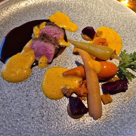 Ein Gourmetgericht mit einem Stück Fleisch, einer roten Sauce, einer gelben Sauce und garniert mit Karotten, Rote Bete und Kräutern auf einem grauen Teller.