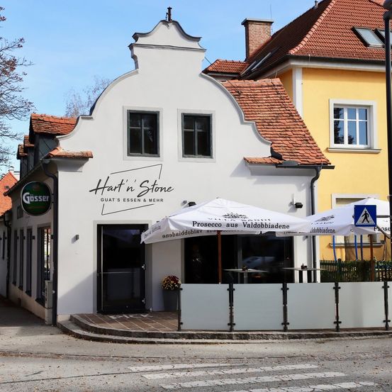 Ein weißes Gebäude mit rotem Dach und einem Schild, das 'Hart n Stone' steht. Es hat zwei Fenster, eine Tür und einen Schirm davor. Das Gebäude befindet sich neben einem gelben Gebäude mit Fenster und Schild.