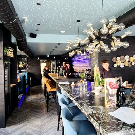 Ein moderner Barbereich mit Marmortheke, blauen Stühlen und einer Kronleuchter. Das Personal steht hinter der Theke, und ein Mann steht in der Nähe.