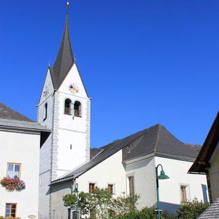 Eine weiße Kirche mit einem Turm und einem Uhrturm steht in einem Dorf unter einem klaren blauen Himmel.