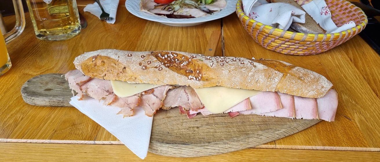 Ein Holztisch zeigt ein langes Sandwich mit Schinken und Käse auf einem Holzbrett, begleitet von einem Korb mit Brot und einer Schüssel Salat.