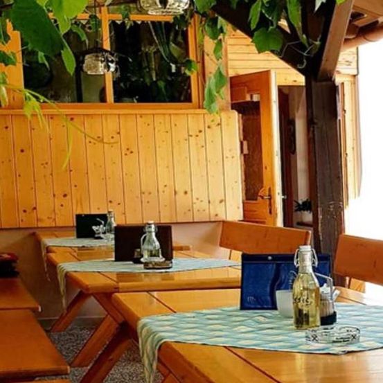 Ein leerer Außenbereich zum Essen mit Holztischen und Bänken. Auf den Tischen stehen Flaschen, eine blaue Tasche und eine Aschenschale. Der Raum hat Holzwände und eine Tür.