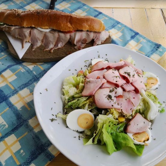 Ein Teller mit Salat, Schinken und Eiern steht auf einem Tisch mit einem Sandwich auf einem Holzbrett.