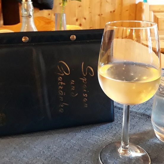 Ein Glas Weißwein steht auf einem Tisch neben einem schwarzen Menü mit goldener Schrift. Hinter dem Menü ist eine Vase mit einer Blume zu sehen.