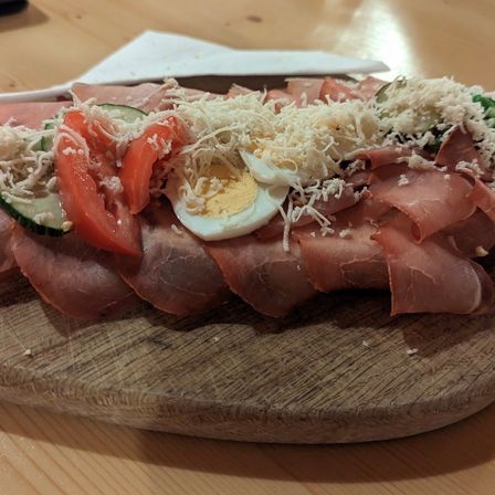 Ein Holzteller mit einem Gericht aus geschnittenem Fleisch, Gurke, Tomate, Ei und geriebenem Käse auf einem Holztisch.