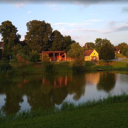 Eine ruhige ländliche Szene mit einem gelben Haus am See, umgeben von Bäumen und Grün. Der Himmel ist klar mit einigen Wolken.