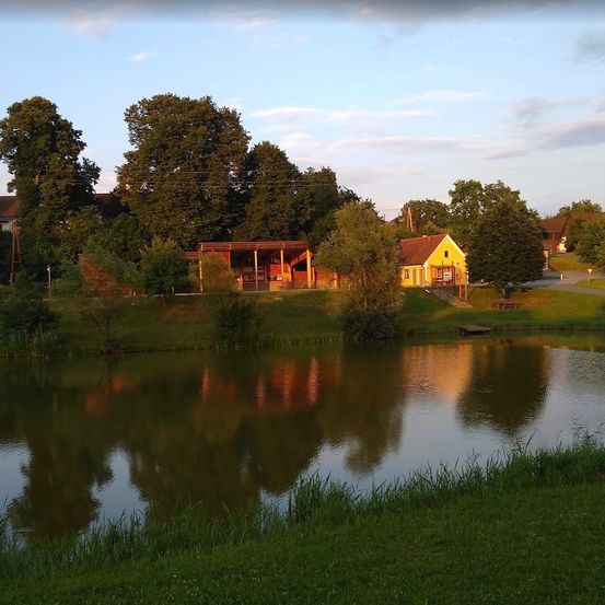Eine ruhige ländliche Szene mit einem gelben Haus am See, umgeben von Bäumen und Grün. Der Himmel ist klar mit einigen Wolken.