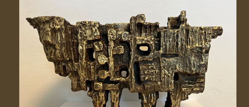 Eine Bronzeskulptur ist auf einer weißen Plattform vor einem braunen Hintergrund ausgestellt. Die Skulptur hat ein komplexes Design mit verschiedenen geometrischen Formen und Öffnungen. Es scheint sich um ein Denkmal für einen Salzburger Künstler zu handeln.