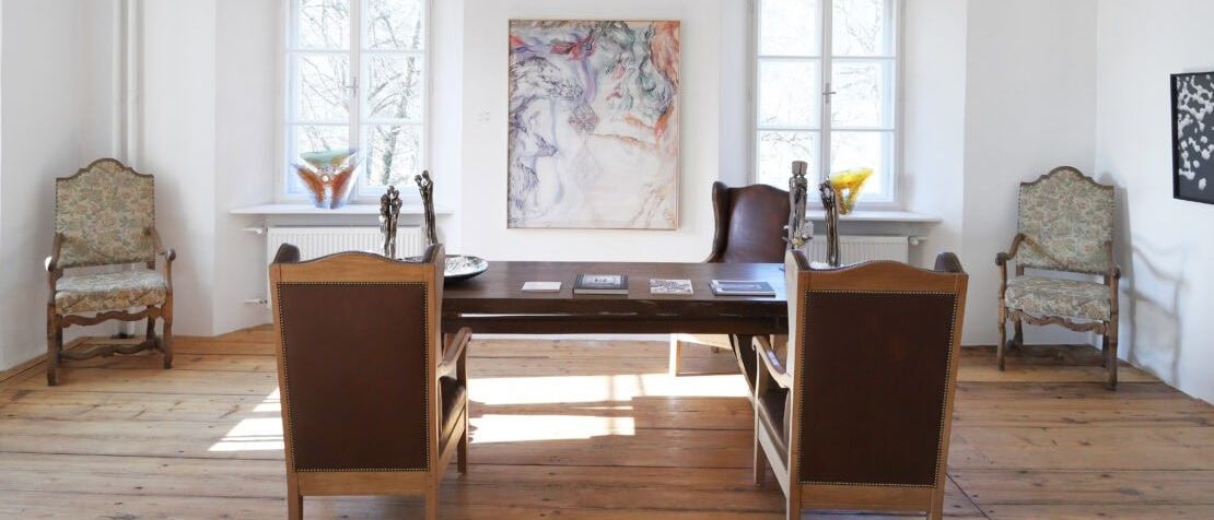 Ein Raum mit Holzboden verfügt über einen Holztisch und zwei braune Lederstühle. Ein großes Gemälde schmückt die Wand über dem Tisch, und eine Kristallkronleuchter hängt von der Decke. Zwei Fenster befinden sich auf beiden Seiten des Gemäldes, mit weißen Rahmen und weißen Heizkörpern darunter.