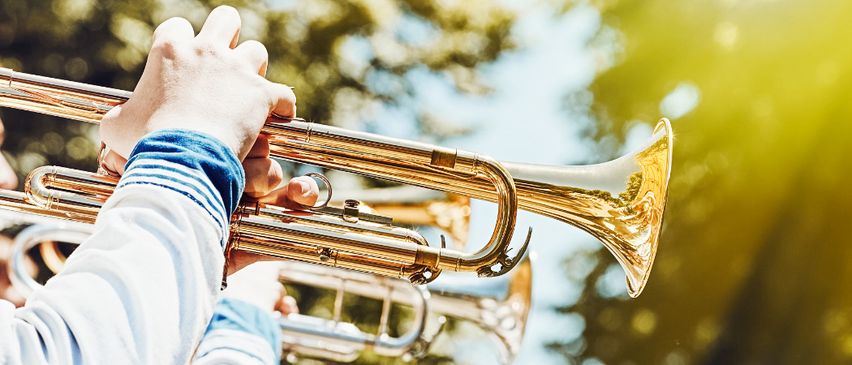 Eine Hand hält eine Messingtrompete im Freien, wobei das Sonnenlicht auf dem Instrument reflektiert und Bäume im Hintergrund sind.