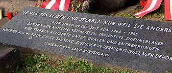 Ein Denkmal mit einem Kreuz und Kränzen steht zwischen großen Felsen. Die Tafel besagt: 'Sie lebten und starben nur, weil sie anders waren.' Blumen und Kerzen umgeben das Denkmal.