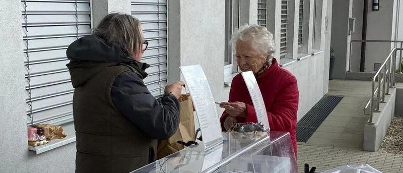 Zwei Frauen stehen vor einem Gebäude, eine hält ein Menü und die andere einen Stift. In der Nähe befindet sich eine Glasvitrine mit Schokoladen.
