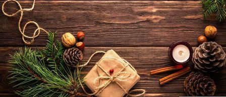 Ein Holztisch zeigt eine Feiertagsszene mit Tannenzapfen, einem verpackten Geschenk, einer brennenden Kerze und einer Feiertagskarte.