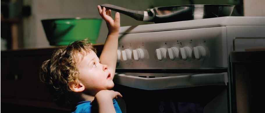 Ein kleines Kind steht auf einem Stuhl und greift nach einer Bratpfanne auf einem Herd. Ein grüner Behälter steht hinter dem Kind auf einem Tisch.