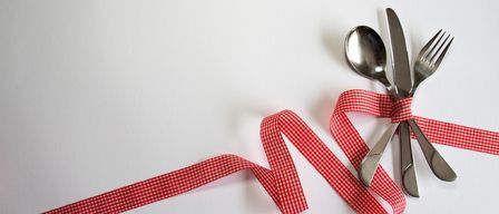 Ein rotes Band mit kariertem Muster ist um einen Löffel, eine Gabel und ein Messer auf einer weißen Oberfläche gebunden.