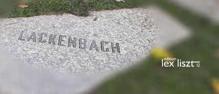 Ein Gedenkstein für 'Lackenbach' ist auf einem Betonweg eingraviert. Im Hintergrund ist eine verschwommene Reflexion eines Gebäudes auf dem Wasser zu sehen.