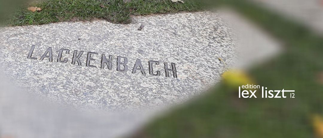 Ein Gedenkstein für 'Lackenbach' ist auf einem Betonweg eingraviert. Im Hintergrund ist eine verschwommene Reflexion eines Gebäudes auf dem Wasser zu sehen.