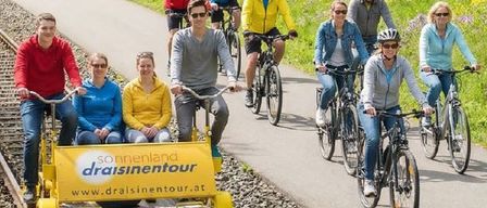 Eine Gruppe von Radfahrern fährt auf einer Bahn, mit Helmen und Fahrradausrüstung, mit einem gelben Wagen mit der Aufschrift 'draisinentour'.