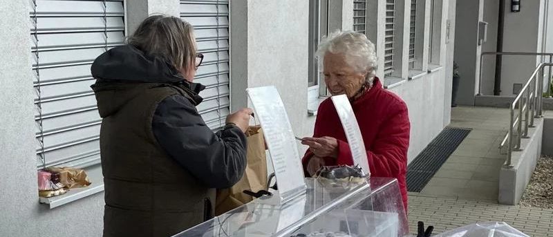 Zwei Frauen, eine mit einem Menü, stehen vor einer Vitrine mit Pralinen, mit Taschen und einem Fenster im Hintergrund.