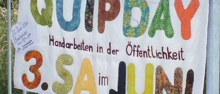 Ein weißes Banner an einem Metallzaun trägt die Aufschrift 'QUIPDAY 3.SA im JUNI.' Darunter steht 'Handarbeiten in der Öffentlichkeit.'