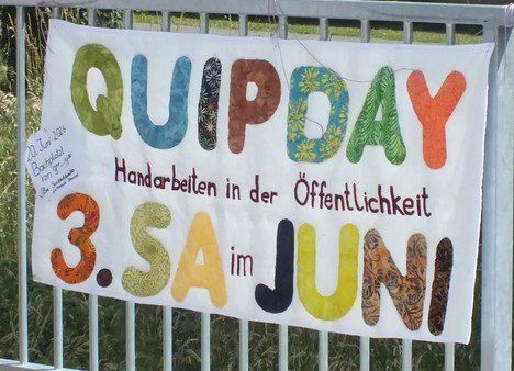 Ein weißes Banner an einem Metallzaun trägt die Aufschrift 'QUIPDAY 3.SA im JUNI.' Darunter steht 'Handarbeiten in der Öffentlichkeit.'