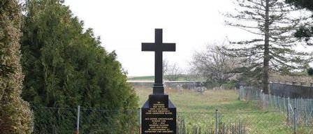 Ein schwarzes Kreuz-Denkmal steht auf einem Rasen, umgeben von einem Zaun. Es hat eine Tafel mit fremdsprachigem Text und ein kleines Blumenarrangement in der Nähe. Bäume und Büsche im Hintergrund.