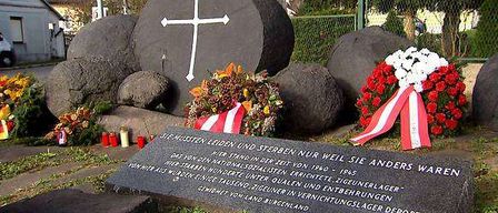 Ein Denkmal mit einem großen Kreuz und Felsen, umgeben von Blumen und Kränzen, mit einer Plakette in Deutsch.