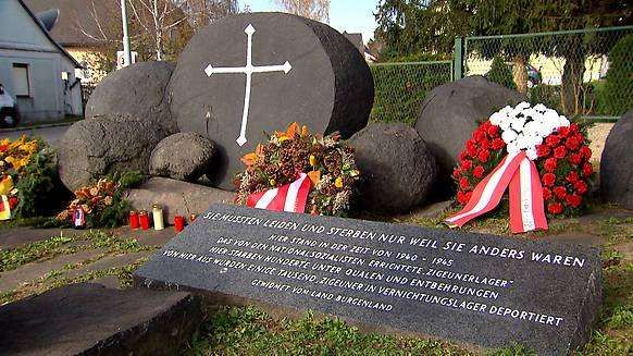 Ein Denkmal mit einem großen Kreuz und Felsen, umgeben von Blumen und Kränzen, mit einer Plakette in Deutsch.