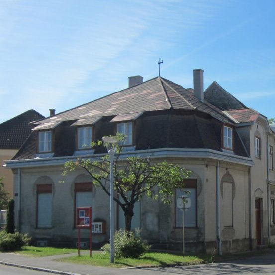 Ein zweistöckiges Gebäude mit einem braunen Dach und mehreren Fenstern, einem Kreuz darauf. Es ist von Grünpflanzen und einer Straßenlampe umgeben.