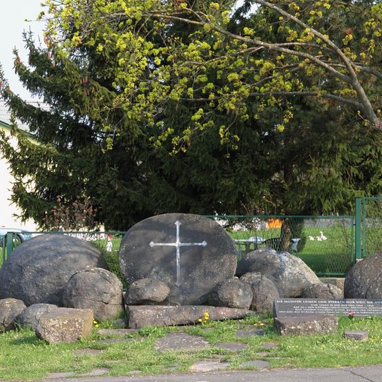 Ein großer Stein mit einem Kreuz in einem Parkbereich, umgeben von anderen Steinen und grünem Gras. Bäume und ein Zaun im Hintergrund.