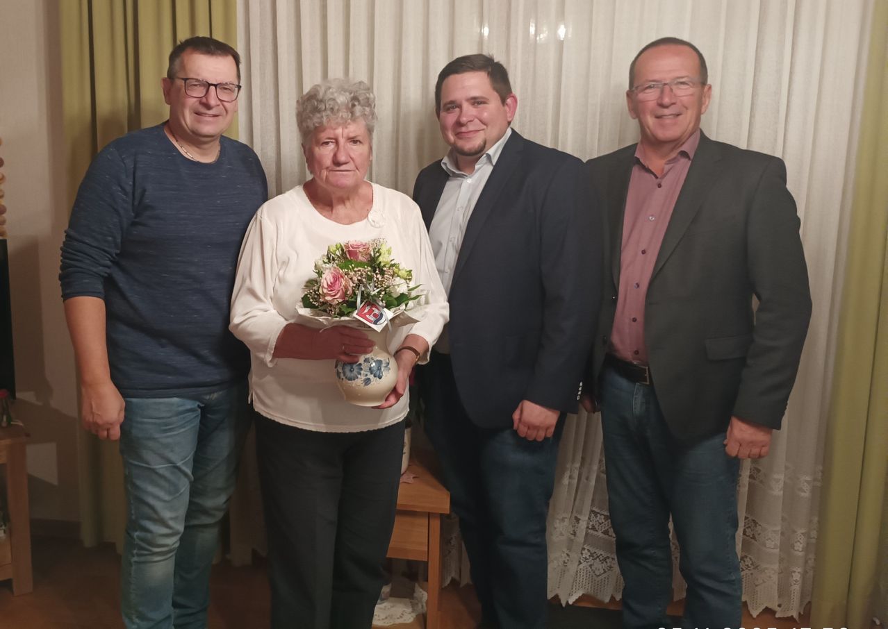 Fünf Personen posieren für ein Foto, wobei eine Frau in der Mitte einen Blumenstrauß in einer Vase hält. Alle lächeln.