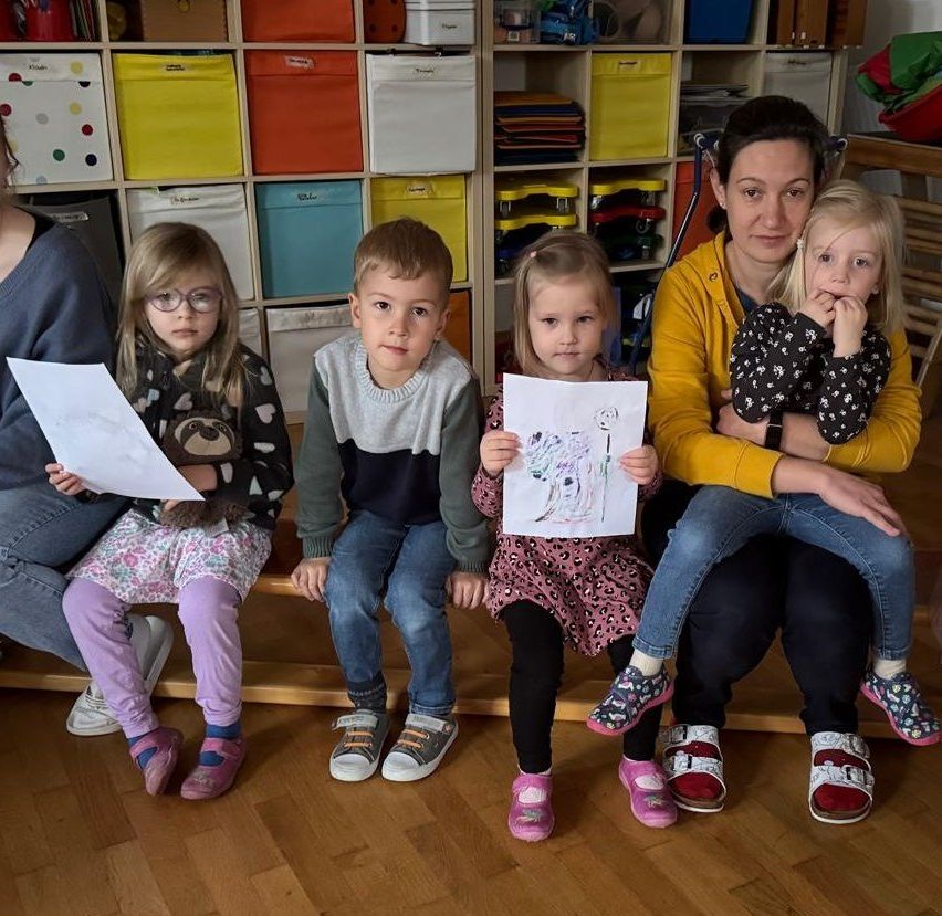 Fünf Kinder und eine erwachsene Frau sitzen auf einer Bank, halten Zeichnungen in der Hand, in einem Raum mit farbenfrohen Aufbewahrungsboxen an Regalen.
