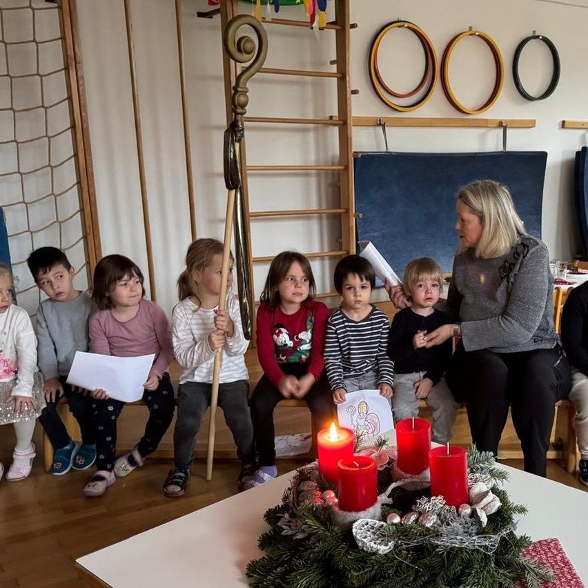 Eine Frau liest einer Gruppe von Kindern eine Geschichte vor, die um einen kerzenbeleuchteten Kranz herumsitzen, mit einem Stab im Vordergrund.