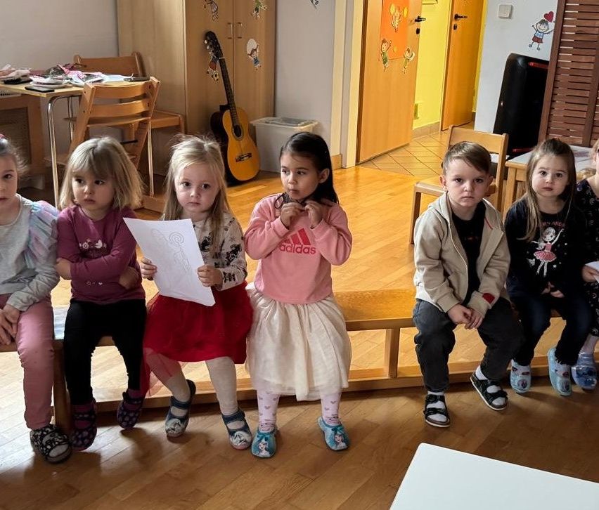 Fünf kleine Kinder sitzen auf einer Bank in einem Klassenzimmer, eines hält ein Papier. Eine Gitarre lehnt an der Wand. Der Raum hat Holzböden und Möbel.