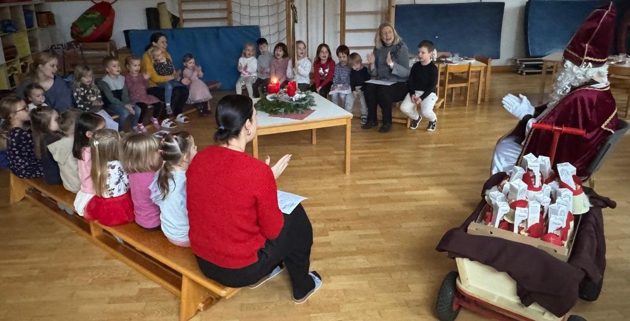 Eine Gruppe von Kindern und Erwachsenen versammelt sich in einem Raum. Eine Frau führt die Gruppe vor einem Tisch mit einer Kerze an.