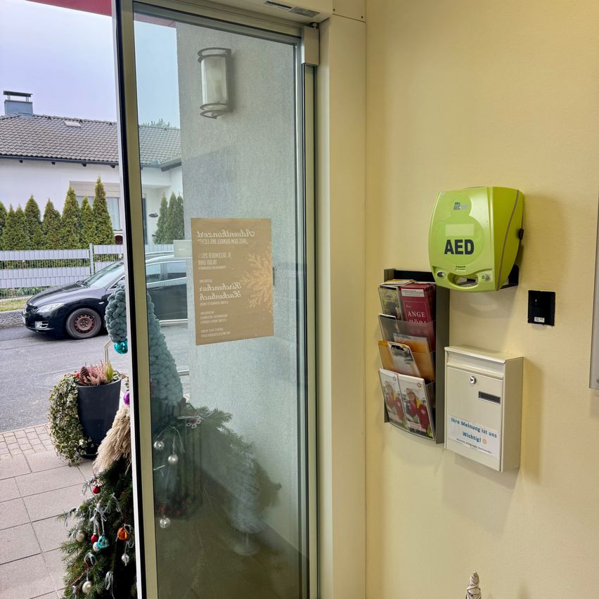 Eine Glastür zeigt einen Flyer an. Ein AED-Gerät und ein Briefkasten sind an der Wand montiert. Ein Auto ist auf der Straße geparkt.