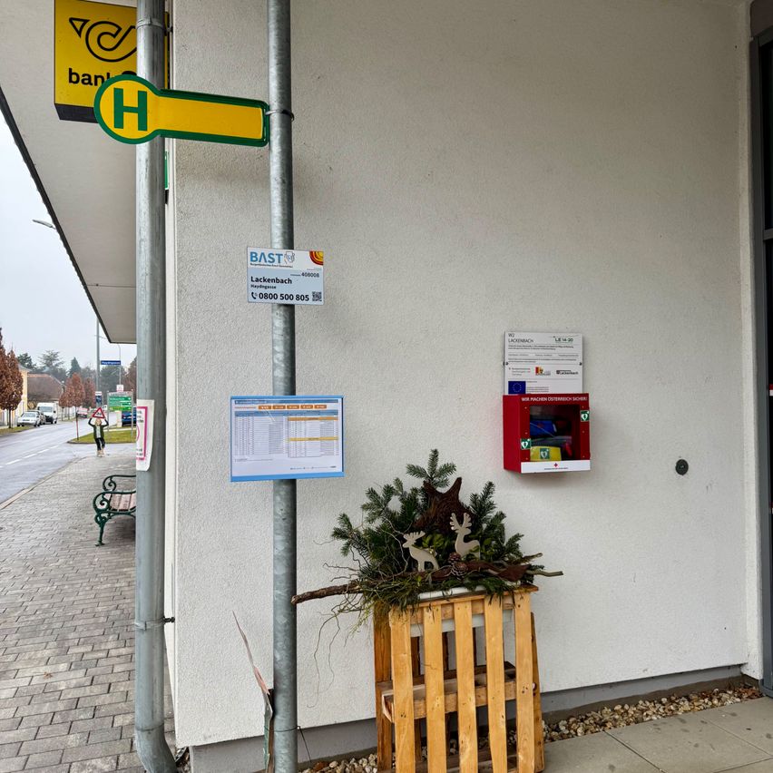 Eine Gebäudewand mit einem Schild für Bast und einer roten Box für Lebensmittelspenden. Ein hölzerner Kasten mit einem Tannenbaum davor steht vor der Wand.