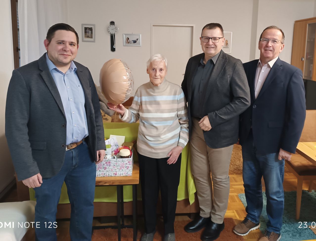 Vier Personen stehen in einem Raum. Eine ältere Frau hält einen Ballon und ein Mann neben ihr einen Geschenkkarton. Dahinter steht ein Tisch mit einem Geschenkkarton und verschiedenen Gegenständen. Gerahmte Bilder und eine Uhr hängen an der Wand.